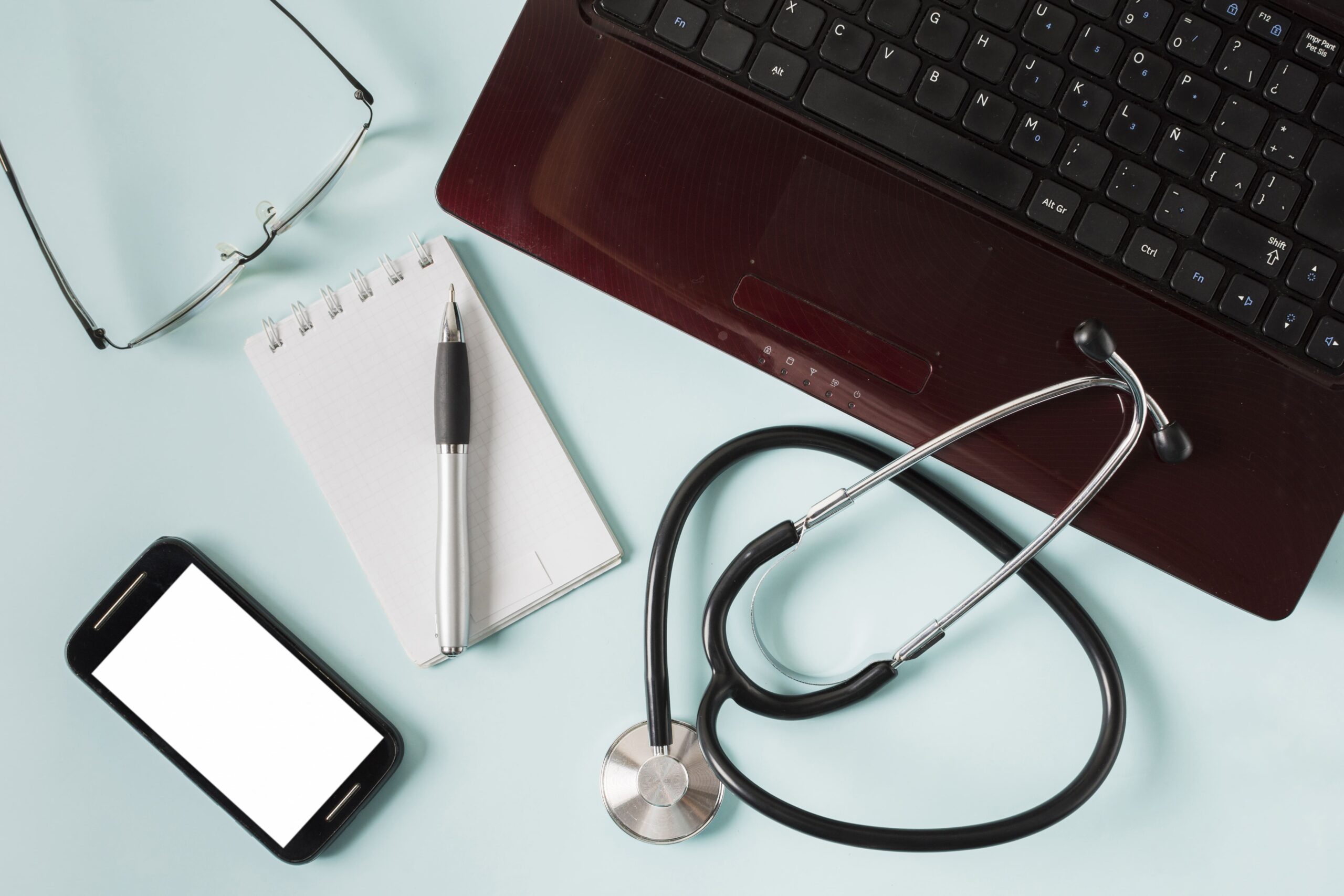 notebook, estetoscópio, celular, óculos e bloco de notas em cima de um fundo azul representando os tipos de software para médicos